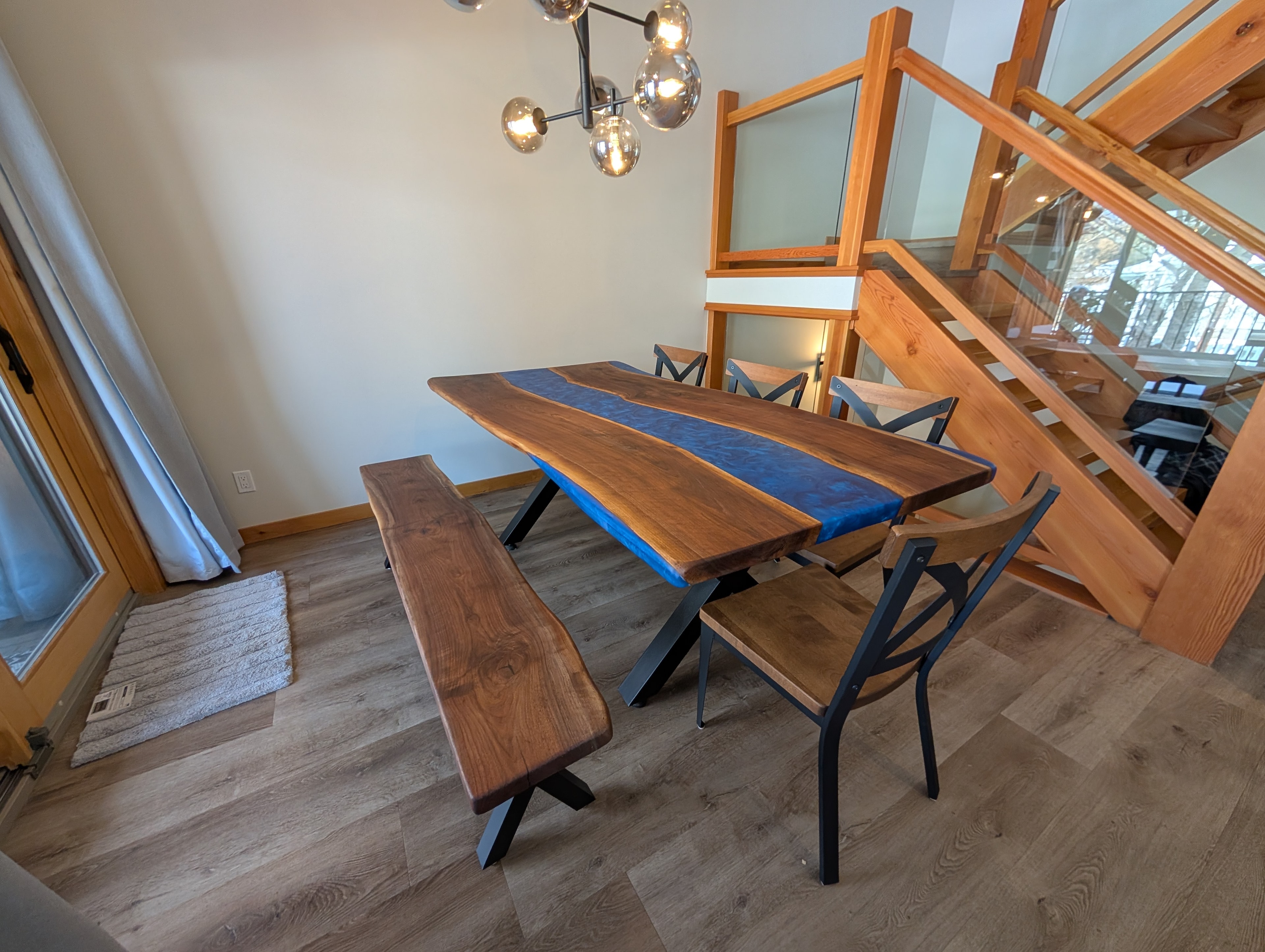 Blue epoxy river walnut dining table with matching bench beside glass staircase
